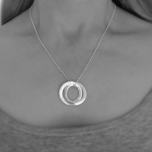 Sterling silver interlocking circle necklace with one hammered and one satin-finish ring on a silver belcher chain, shown on a model wearing a simple top.