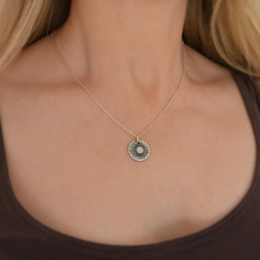 Handmade sterling silver hammered disc necklace with a textured and oxidised finish, featuring a small central disc on a silver trace chain, shown on a model wearing a simple top.